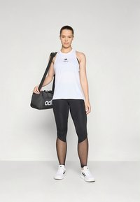 Valkoinen hihaton urheilutoppi, jossa musta logo, yhdistetty mustiin legginsseihin, joissa on läpikuultavia paneeleja, ja musta matkalaukku, jota malli pitää kädessään.