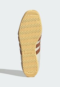 Brown and white sneaker with a textured rubber sole featuring a zigzag pattern, and a visible logo. The upper is designed with stripes.