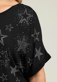 Czarna bluzka z krótkim rękawem, ozdobiona srebrnymi wzorami gwiazd z kryształków w różnych rozmiarach oraz rozproszonymi ozdobami, o luźnym kroju i miękkiej tkaninie.