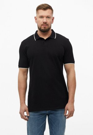 Mann trägt ein schwarzes kurzärmeliges Poloshirt mit weißem Rand und blaue Jeans, steht vor einem schlichten weißen Hintergrund.