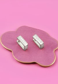 Silvriga örhängen med rektangulär design, prydda med klara stenar monterade i en rad. Liggande på en strukturerad rosa visningsplatta med scallopade kanter.