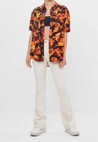 Svart kortärmad skjorta med ett eldig orange och gul eld-mönster, bärs över en svart crop top, kombineras med vita utsvängda byxor och vita sneakers.