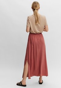 Maxi-rok in roestkleur met zijsplitten, gemaakt van een gladde stof. Gecombineerd met een lichtbeige top, gedragen met zwarte sandalen.