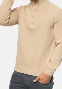 Beige gebreide trui met een hoge kraag, voorzien van een knoopsluiting met vier knopen, geribde manchetten en zoom. Gladde textuur en aansluitende pasvorm.