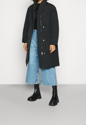 Sort lang frakke med høj krave, trykknaps- og lynlås-lukkninger, parret med lyseblå brede jeans og sorte chunky støvler.