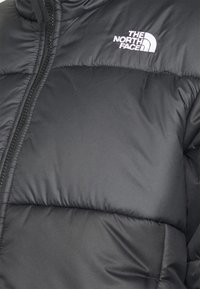 Veste matelassée noire avec design matelassé, fermeture éclair et logo brodé. Texture lisse avec rembourrage isolant pour la chaleur.