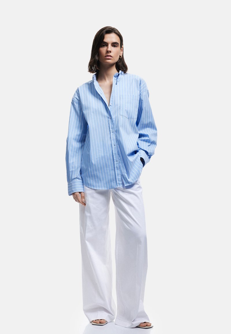 Donna che indossa una camicia blu a righe larghe e pantaloni bianchi larghi, in piedi con una mano in tasca contro uno sfondo neutro.