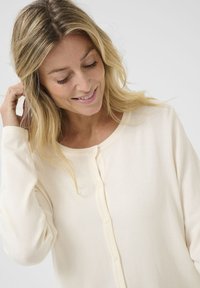 Cardigan beige chiaro con maniche lunghe, scollatura rotonda e chiusura a cinque bottoni. Tessuto morbido con una finitura liscia; design minimalista.