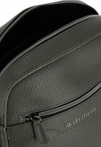 Sac à dos en cuir vert avec une finition texturée, comportant une fermeture éclair, des coutures visibles et un accent logo.