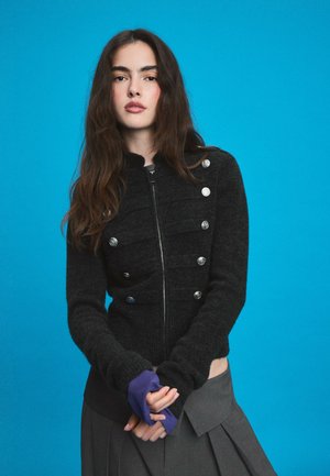 Jeune femme aux longs cheveux bruns portant une veste noire courte, un haut violet et un pantalon large gris plissé sur fond bleu.