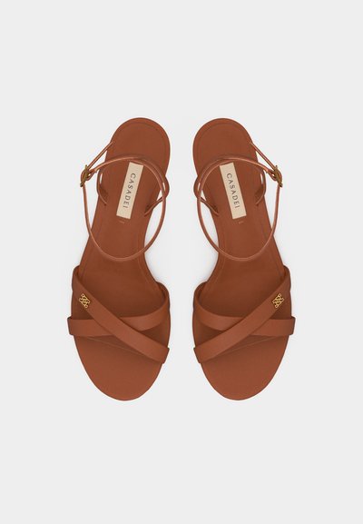 Καφέ δερμάτινα σανδάλια με λουριά που διασταυρώνονται, αγκράφες στον αστράγαλο και χρυσό λογότυπο της επωνυμίας σε καθαρό λευκό φόντο.