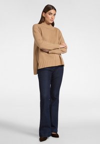 Beige stickad tröja med hög hals, ribbat kant och sidofyra, tillsammans med mörka utsvängda jeans och bruna spetsiga skor.