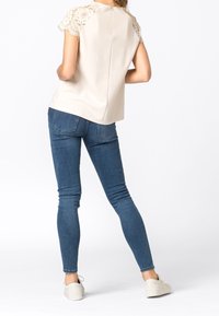 Beige top met kanten schouderaccenten, gecombineerd met donkerblauwe skinny jeans en lichtgekleurde sneakers, gezien vanaf de achterkant.