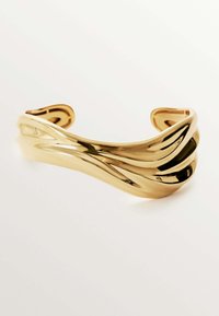 Pulsera de puño en tono dorado con un diseño ondulado, superficie lisa y una estructura abierta, que muestra una estética moderna y escultórica.