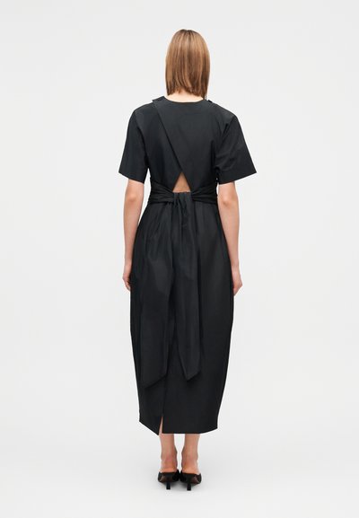 Woman standing facing away wearing a black midi dress with short sleeves, a small back cutout, and tied waist detail, paired with black heels.