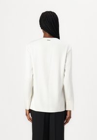 Blusa bianca a maniche lunghe in tessuto liscio con scollo rotondo, fessure laterali e una targhetta metallica sul retro. Abbinata a pantaloni neri.