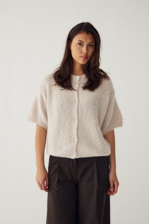 Jeune femme aux longs cheveux foncés porte un pull beige clair à manches courtes boutonné et un pantalon large foncé, debout devant un fond uni.