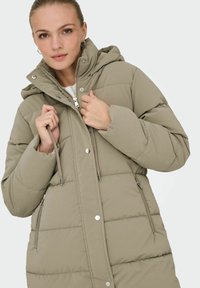 Polstret parka-jakke i lys olivengrøn med aftagelig hætte, lynlås og trykknapper samt to sidelommer. Blød, quiltet tekstur.