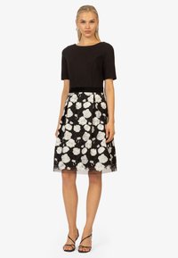 Schwarzes Kleid mit einem taillierten Oberteil und einem ausgestellten Rock, der mit weißer Blumenstickerei verziert ist. Der Stoff hat eine glatte Textur, mit einer durchsichtigen Überlagerung am Saum.