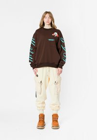 Sudadera marrón con texto y gráfico en turquesa, combinada con pantalones cargo color crema que tienen bolsillos con cremallera y botas marrones con cordones.