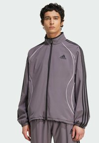 Siva športna jakna z črnimi črtami, belim obrobljanjem, z zadrgo spredaj in elastičnimi manšetami. Ima adidas logotip na levem prsnem delu.