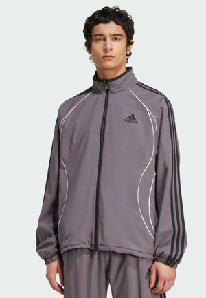 Szürke sportdzseki fekete csíkokkal, fehér díszítéssel, cipzáras elülső résszel és rugalmas mandzsettákkal. Az adidas logója a bal mellen található.