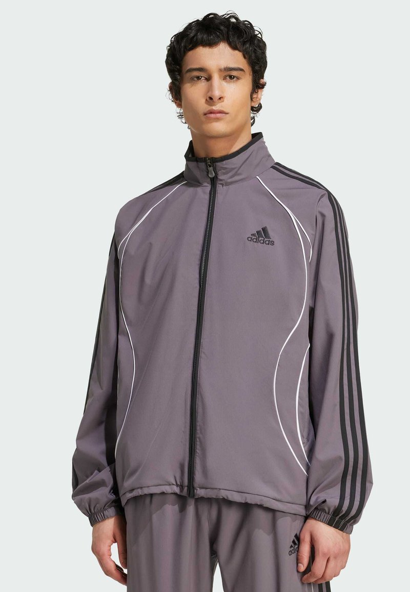 Siva športna jakna z črnimi črtami, belim obrobljanjem, z zadrgo spredaj in elastičnimi manšetami. Ima adidas logotip na levem prsnem delu.