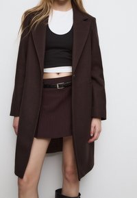 Cappotto in misto lana marrone con design sartoriale, maniche lunghe e spacchi laterali. Sotto, un top corto nero e una gonna bordeaux a righe con una cintura.