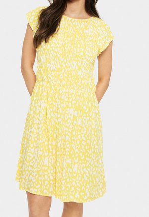 Woman wearing a yellow and white patterned short-sleeve dress with a gathered waist and knee-length skirt, standing with hands in pockets.