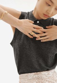 Top sin mangas de color gris oscuro con cuello, combinado con una falda ligera con lentejuelas. Manos adornadas con joyas de oro, incluyendo anillos y pulseras.