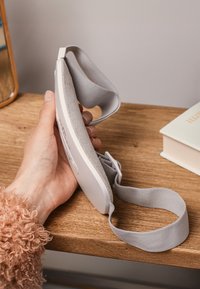 Weißer ergonomischer Augenschutz mit einem weichen grauen Riemen, der eine glatte Oberfläche und eine plüschige Unterseite aufweist, gegen einen Holztisch gehalten.