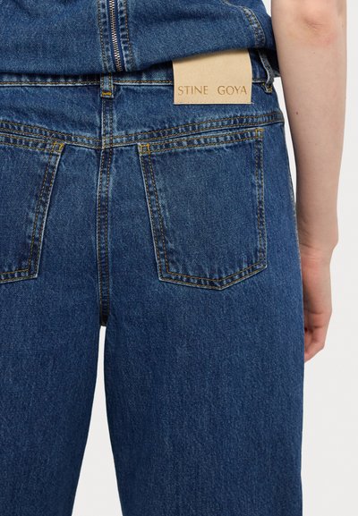 Jean en denim bleu à taille haute, avec deux poches arrière, des coutures visibles et une étiquette en cuir portant la marque "STINE GOYA" au niveau de la taille.