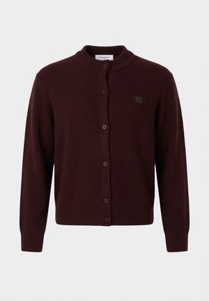 Femme portant une casquette beige, un cardigan marron foncé boutonné, un pantalon ample marron clair et des ballerines noires, debout devant un fond uni.