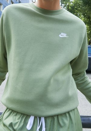 Osoba ubrana w jasnozieloną bluzę Nike i pasujące spodnie z białym sznurowadłem, stojąca na zewnątrz w pobliżu budynków i drzew.