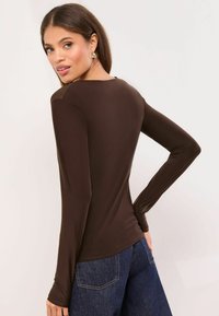 Pruun pikkade varrukatega top, millel on sile ja veniv tekstuur ning ümmargune kaelus; ühendatud tumedate teksadega.