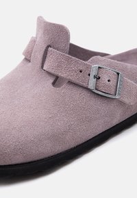 Κοντινό πλάνο ενός μώβ παπουτσιού από σουέντ με ρυθμιζόμενο λουρί και ασημένια πόρπη πάνω σε μαύρη σόλα, σε άσπρο φόντο.