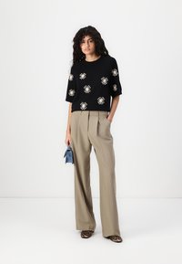 Maglione nero a maniche corte con ricami floreali bianchi, abbinato a pantaloni beige a gamba larga e scarpe con stampa leopardata. Tiene una borsa blu.