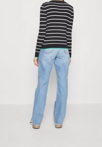Gestreifter schwarz-weißer Pullover mit grünen Akzenten, kombiniert mit hellblauen weit geschnittenen Jeans. Details sind Taschen auf der Rückseite und ein fransiger Saum.