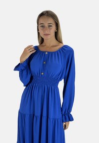 Blå langærmet kjole med en smocket overdel, med firkantet halsudskæring, tre træknapper og flæset nederdel. Blødt, struktureret stof.