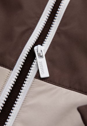 Close-up van een witte rits met "OMBRE" op het trekkerlabel, bevestigd aan stoffen delen in donkerbruine en beige tinten.