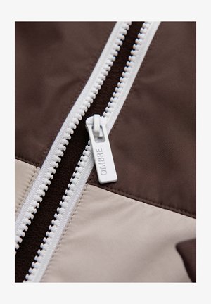 Close-up van een witte rits met "OMBRE" op het trekkerlabel, bevestigd aan stoffen delen in donkerbruine en beige tinten.