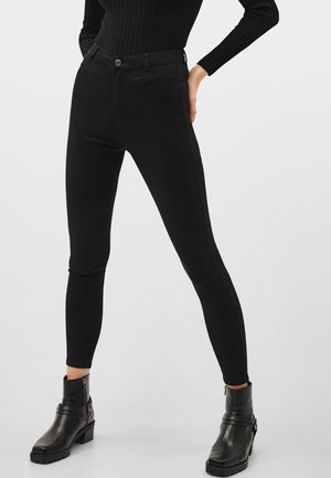 Femme portant un pantalon skinny noir taille haute, un top noir côtelé à manches longues et des bottines noires avec des fermetures éclair sur les côtés et des talons bas.