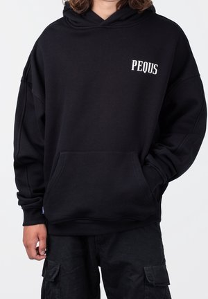 Person trägt einen schwarzen Hoodie mit weißem "PEQUS" auf der linken Brust und schwarze Cargohosen, eine Hand in der Hoodie-Tasche.