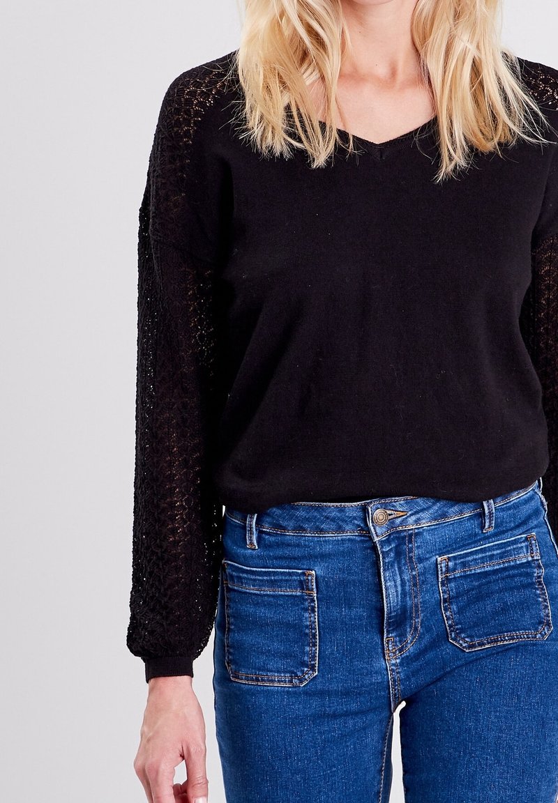 Femme portant un haut noir à manches longues en dentelle et un jean taille haute bleu avec poches plaquées à l'avant, sur un fond uni.