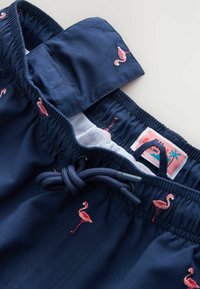 Tengerészkék úszóshort elasztikus derékkal, rózsaszín flamingó hímzéssel és zsinórral. Hálós bélés és márkacímke látható a tetején.