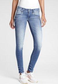 Slim-fit modré džíny nosené s bílými a růžovými teniskami, viditelné od pasu po nohy, které má na sobě stojící osoba na jednobarevném pozadí.