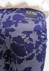 Blå mönstrade jeans med blommönster, texturerad tyg och ett silveremblem på bakfickan med en märkesetikett ovanför.