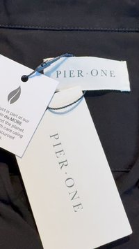 Tecido preto com um rótulo "PIER ONE" anexado. Etiqueta branca com um logótipo de folha e uma mensagem de sustentabilidade. Design simples, sem texturas notáveis.