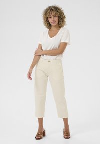 Helle beige, knöchellange Hose aus Baumwollmischung, mit geradem Schnitt und einem goldenen Knopfverschluss. Kombiniert mit einem cremigen V-Ausschnitt-T-Shirt.