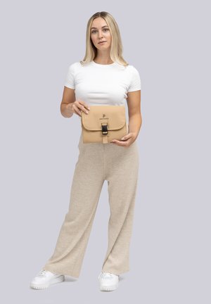 Vrouw in een wit shirt en beige broek houdt een opgevouwen beige tas vast met het "pactastic" logo op een lichtgrijze achtergrond.